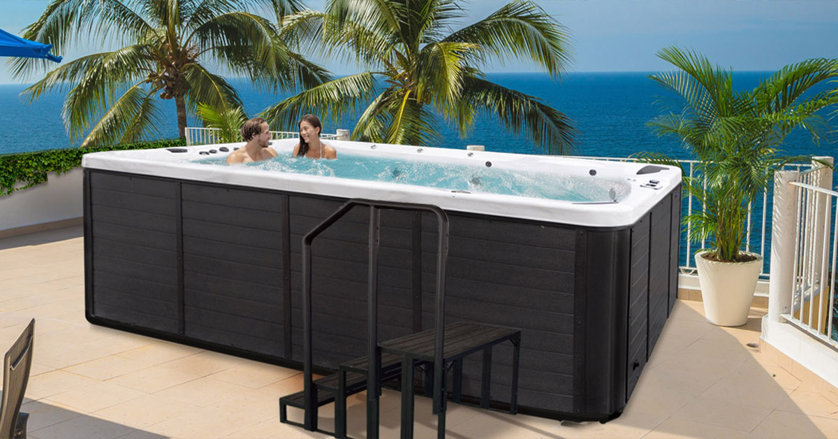 Two people laughing in a Cal Spas Swim Pro™ hot tub, with steps, on a patio with palm trees and ocean in the background.